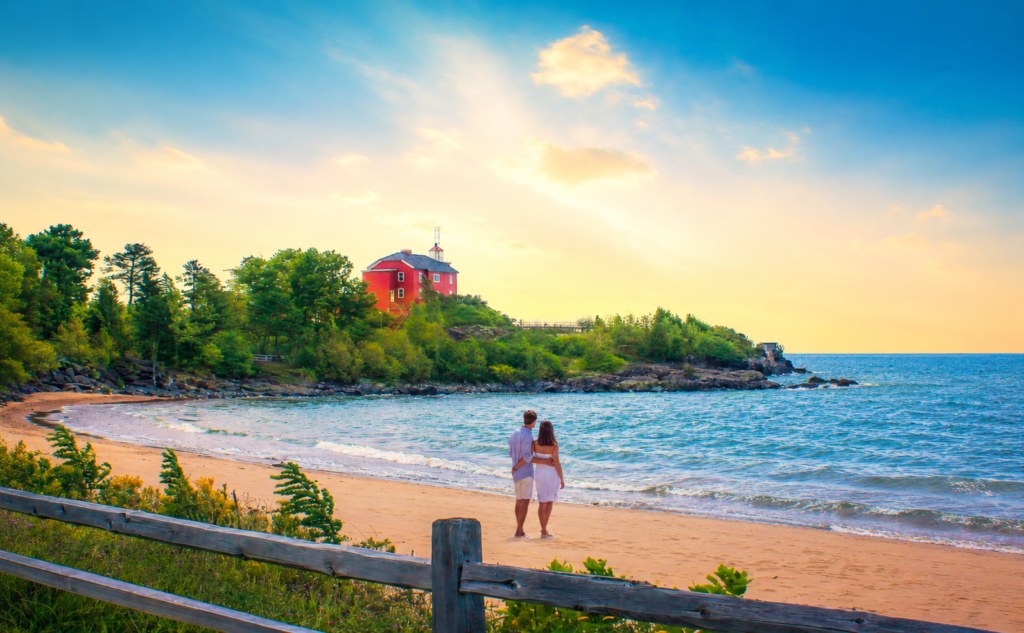 Couple standing on a sunny Lake Superior beach with a red lighthouse in the distance